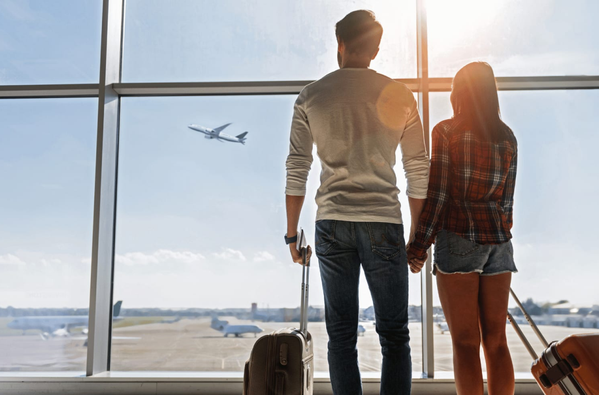 Vue de dos d'un couple à l'aéroport avec des valises, se tenant la main et regardant les avions décoller depuis la salle d'embarquement.