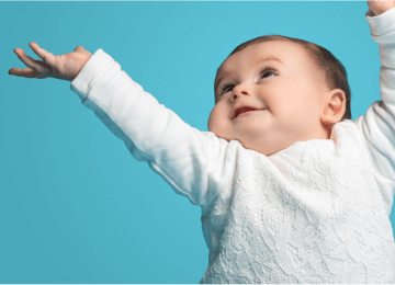 Un bébé en tenue blanche levant les bras avec un sourire joyeux, sur un fond bleu turquoise.