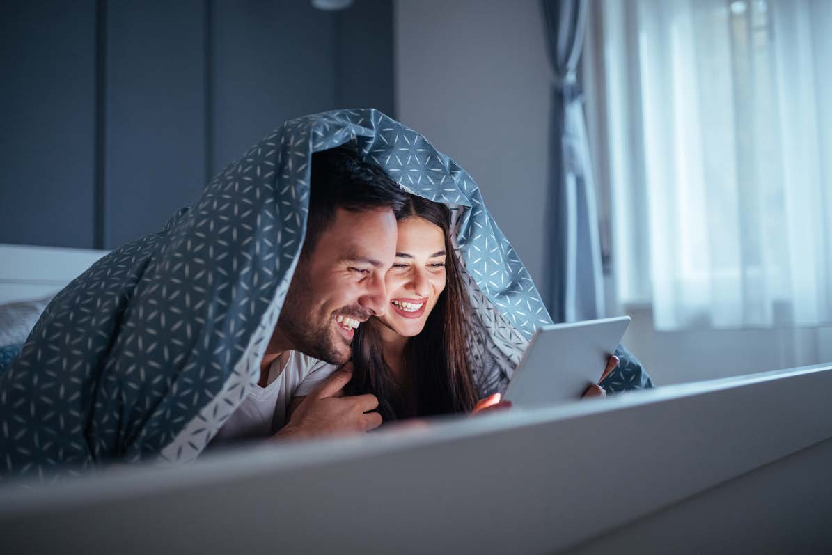 Couple dans une chambre allongé à plat ventre sur leur lit, la couette remontée sur leur tête, regardant un contenu sur leur tablette.