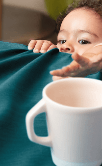 Bébé étendant le bras pour atteindre une théière sur une table recouverte d'une nappe bleue.