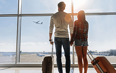 Vue de dos d'un couple à l'aéroport avec des valises, se tenant la main et regardant les avions décoller depuis la salle d'embarquement.